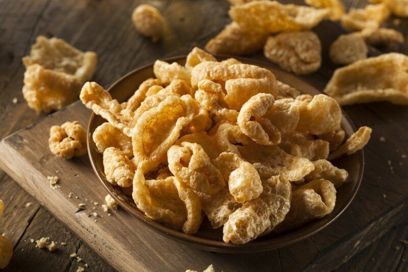 A wooden board with a brown bowl of pork rinds that could be a keto snack