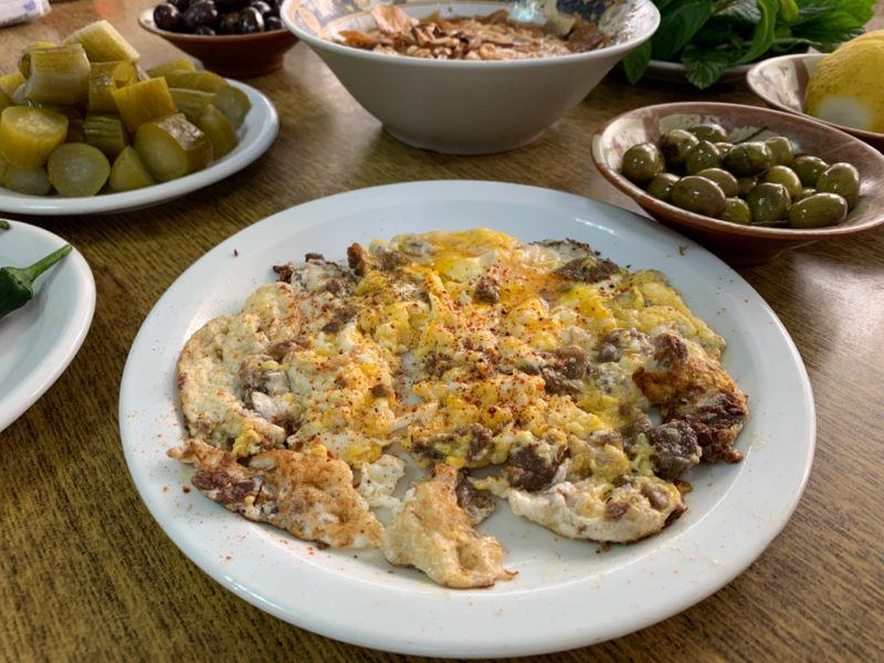 A white plate of scrambled eggs that have been cooked with awarma and other Middle Eastern dishes