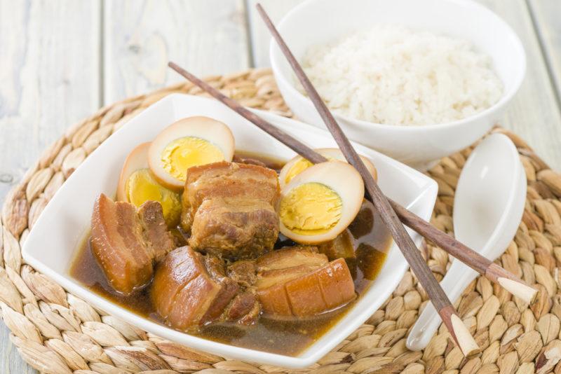 A small white dish filled with pork belly, and an egg, next to chopsticks and an egg