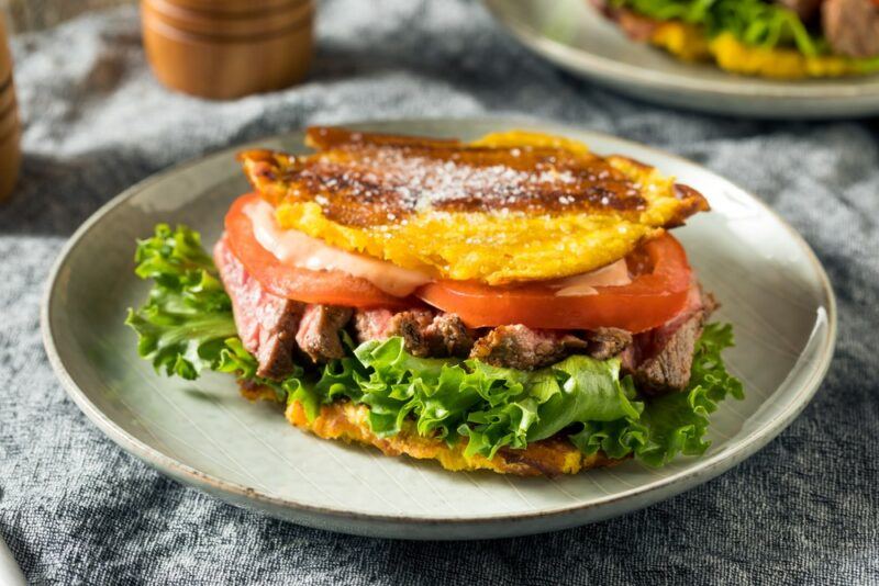 A white dish that contains a plantain-based sandwich called jibarito that includes ingredients like tomatoes and lettuce