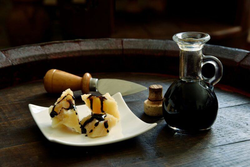 The top of a barrel with a glass jug of balsamic vinegar, plus a white plate of cheese covered in the vinegar