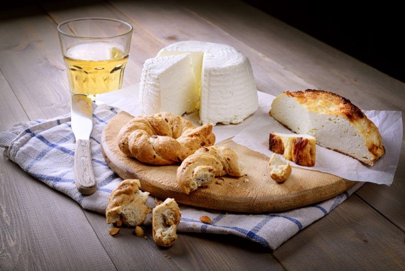 A wooden board on a tea towl with fresh cheese, wine, and bread