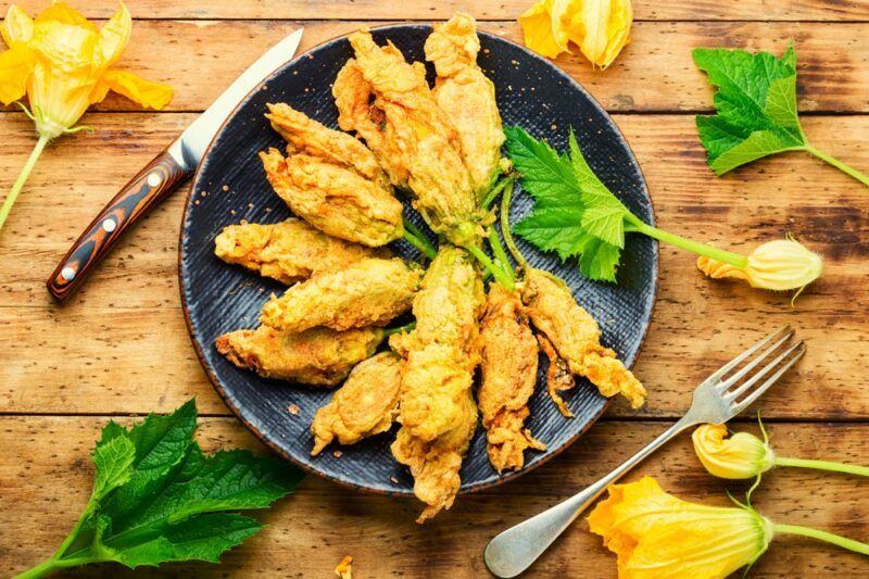 A black dish containing fried zucchini flowers, next to a knife and some flowers scattered across the table