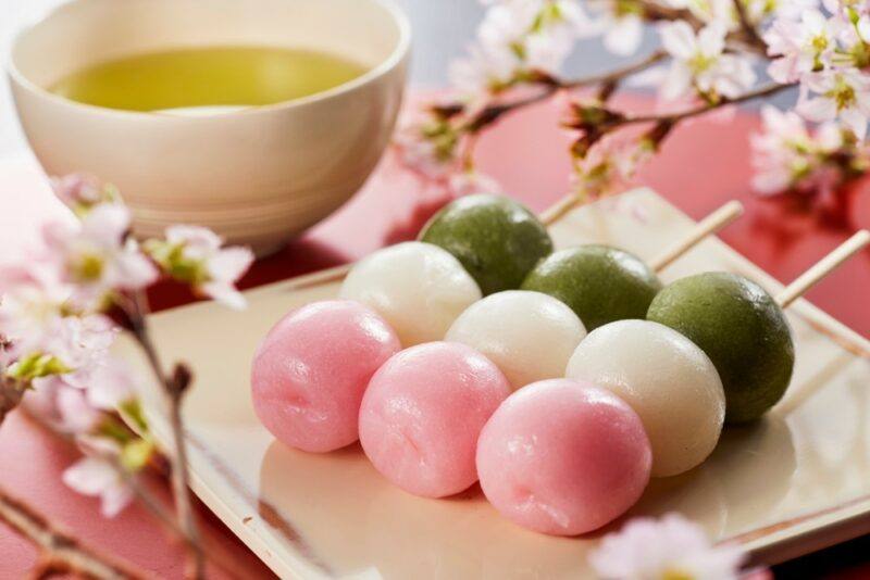 Three skewers each with a green, white, and pink dango rice ball, with flowers in the background and a bowl of sauce or oil