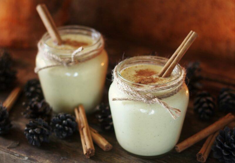 Two jars containing a ponche a creme cocktail, garnished with cinnamon sticks, next to some pine cones