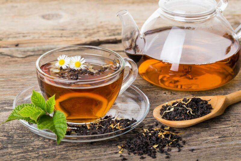 A glass pot of black tea next to a spoon of the tea and a glass mug of brewed green tea