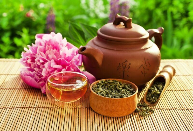 A brown tea pot next to a mug of green tea and a container of green tea leaves, outside on a bamboo mat