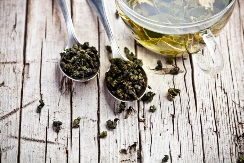 A white table with two spoons of green tea leaves, in front of a pot of green tea