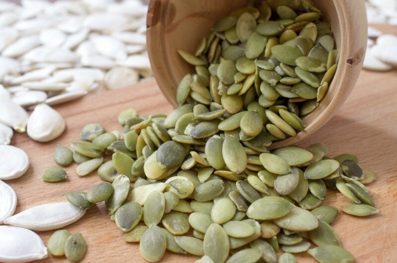 A pot containing green pumpkin seeds spilling out onto a table, surrounded by white pumpkin seeds