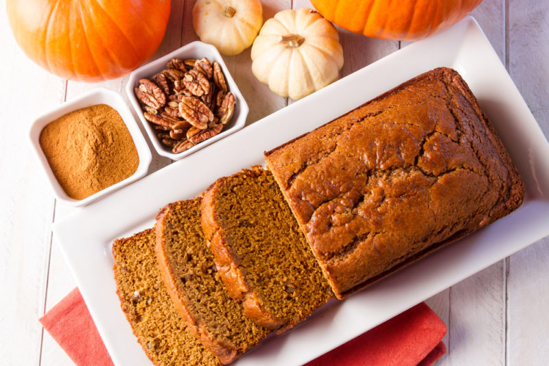 A white plate with a pumpkin loaf that has been sliced next to pumpkin spice and walnuts