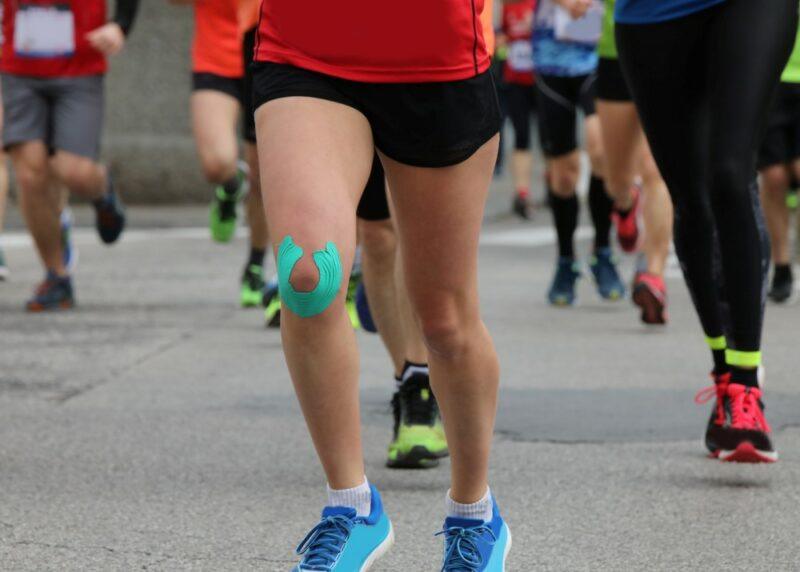 A runner that's part of a race with a red shirt, black shorts, blue shoes, and green tape around one knee