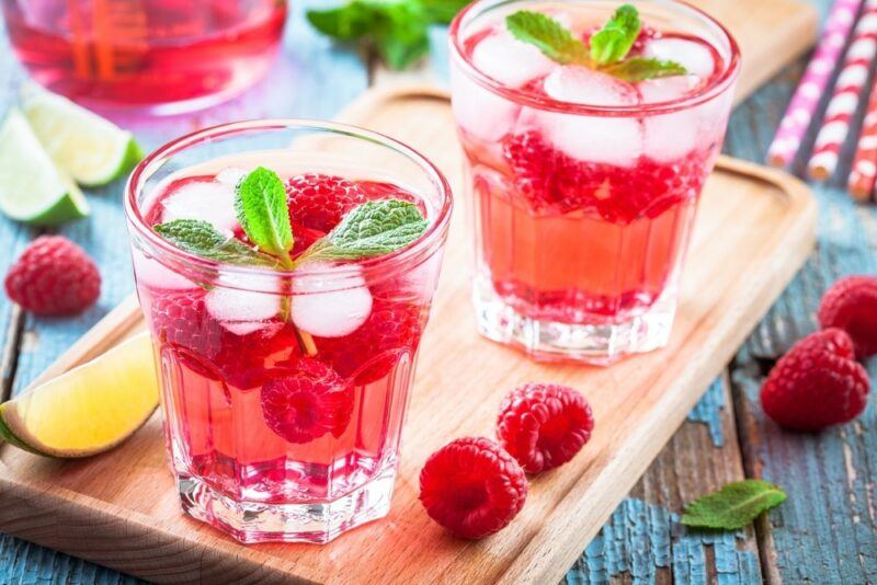 A wooden board with two glasses of a raspberry White Claw cocktail with ice and mint leaves, with some raspberries, lemon, and lime wedges scattered around