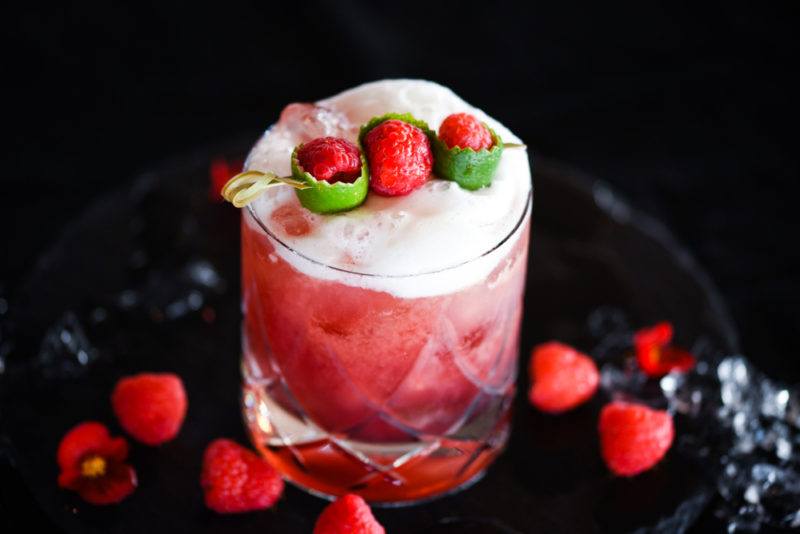 A raspberry cocktail with egg white foam where there are more raspberries on the table and as a garnish