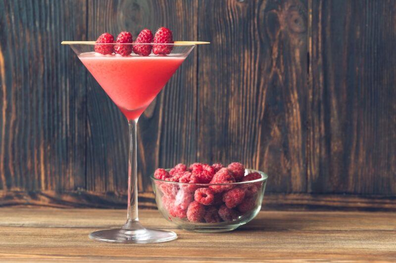 A raspberry martini called a razzitini, garnished with raspberries, next to a dish of raspberries