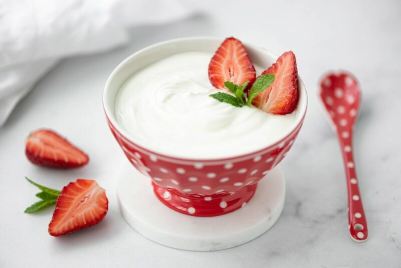 A red and white bowl with spots filled with Greek yogurt and garnished with two strawberry halves, next to a red and white dotted spoon and two more strawberry halves