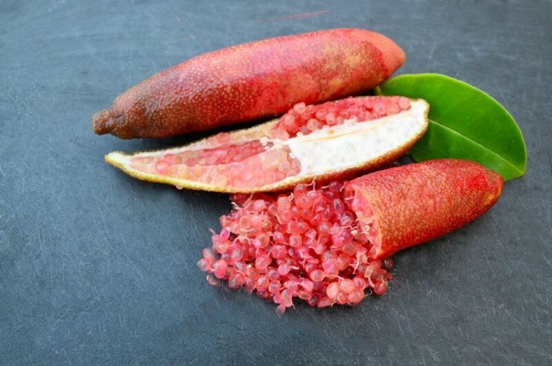 A whole red finger lime, next to half a finger lime and some of the fruit pieces next to it.