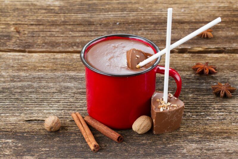 A red mug containing a red wine hot chocolate with chocolate stirrers, with some spices on the table