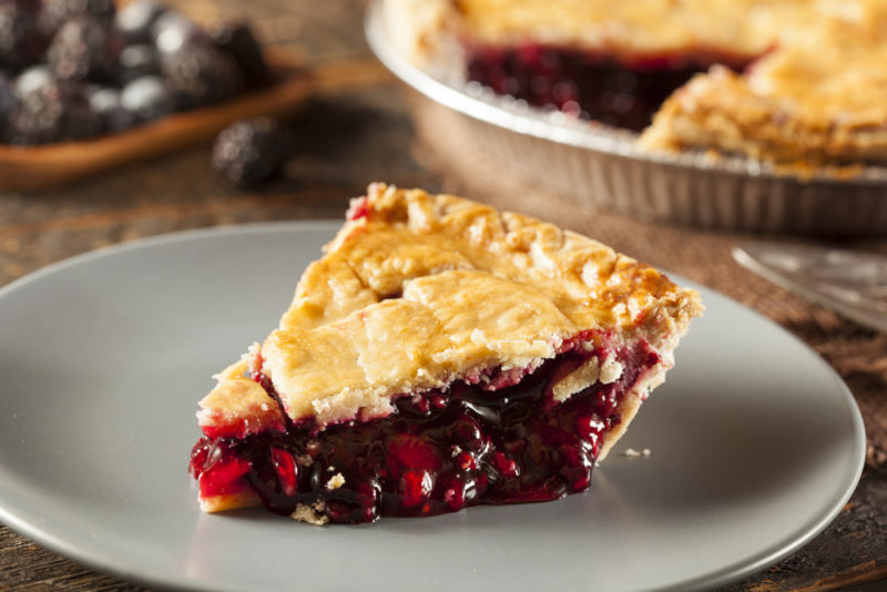 A gray plate with a piece of berry pie, with the rest of the pie in the background