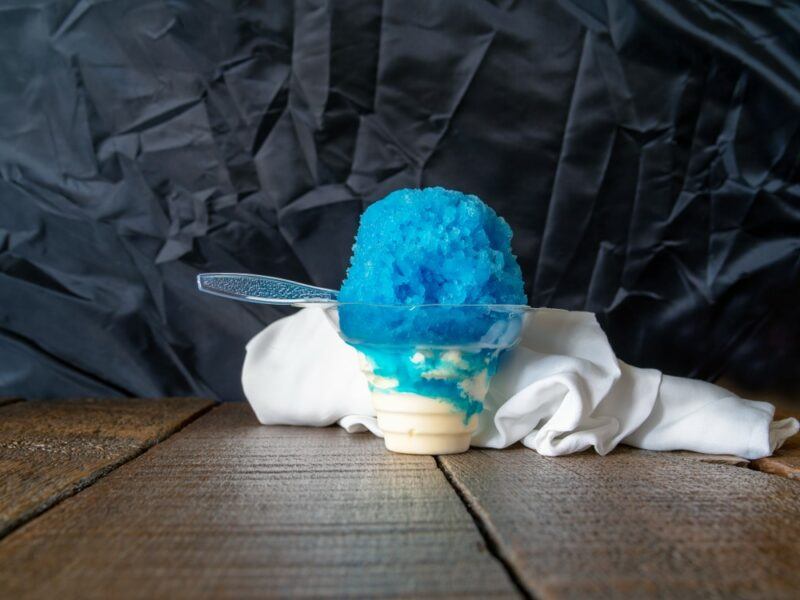 A wooden table with a bright blue rock n roll snow cone that has been drenched in plenty of syrup