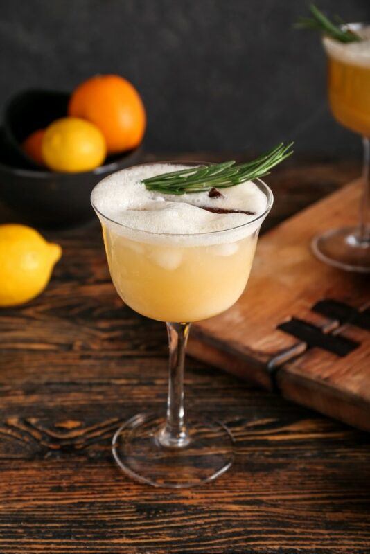 A rosemary sour cocktail on a wooden table, with some lemons in the background