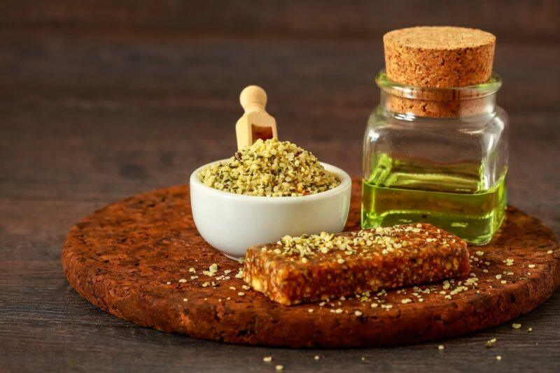 A wooden board with a bowl of hemp hearts, a jar of hemp oil, and a hemp seed protein bar