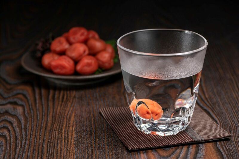 A glass containing a single dried plum and a sake martini, with a plate of plums in the background