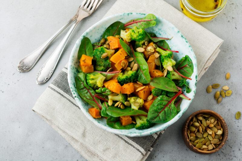 A salad in a white bowl that contains pumpkin and pumpkin seeds, with a small bowl of the seeds next to it