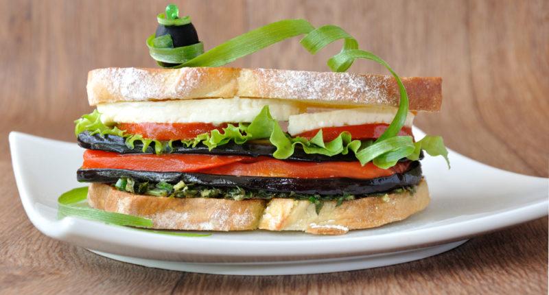 A white plate with an eggplant, cheese, lettuce and tomato sandwich