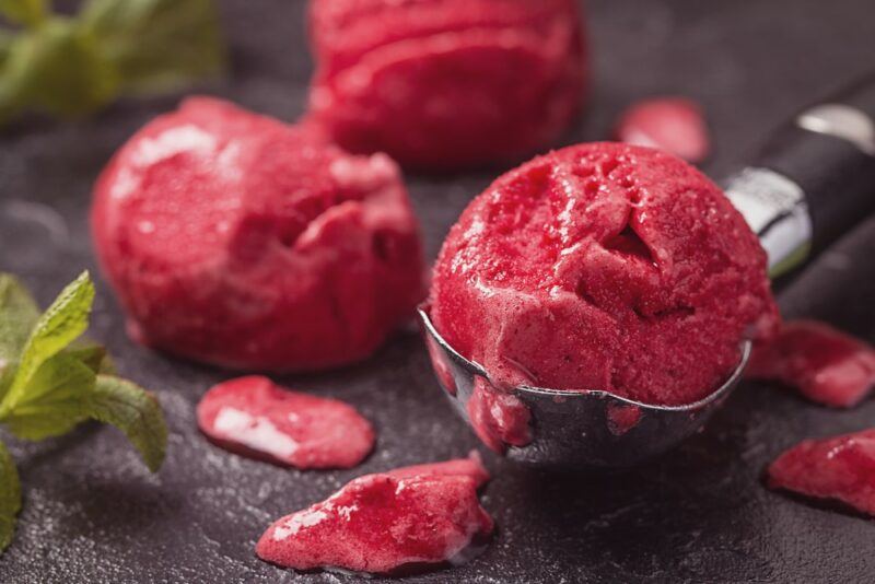 An ice cream scoop with a scoop of berry sorbet, plus two berry sorbet scoops in the background