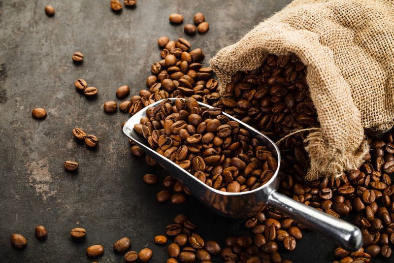 A burlap sack with coffee beans spilling out onto the table. There is a metal scoop with more coffee beans.