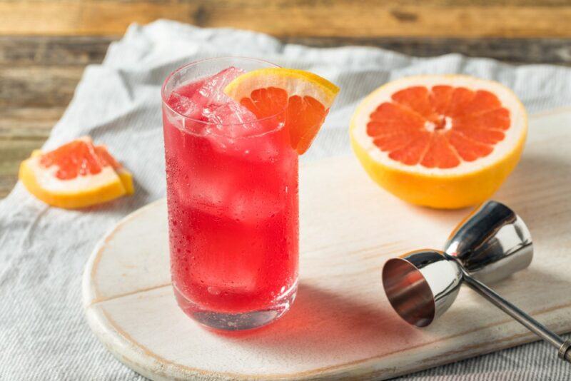 A wooden board with a glass containing a sea breeze cocktail, next to a cocktail jigger and some grapefruit