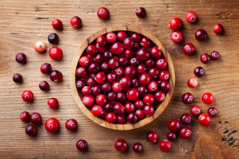 A large wooden bowl of cranberries, with more cranberries scattered across the wooden table