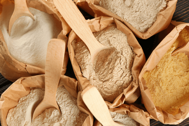 Various paper bags containing different types of gluten free flour, each with a wooden spoon.