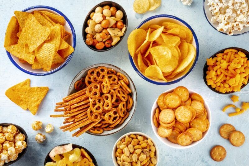 A light colored table with bowls of salty snacks, including chips, nuts, crackers, and pretzels.