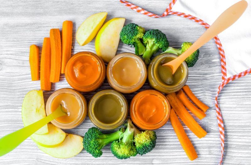 Six glass containers of baby food puree surrounded by vegetables