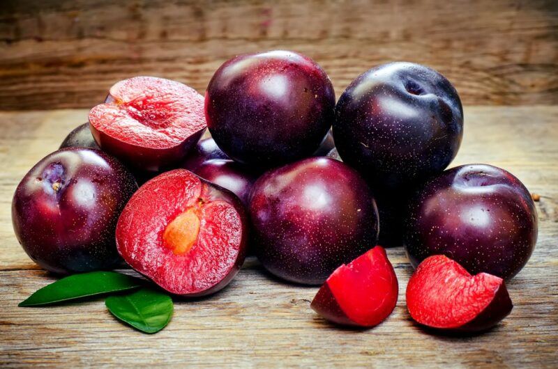 Fresh plums in a pile on a table. A couple have been cut, so their bright interiors can be seen