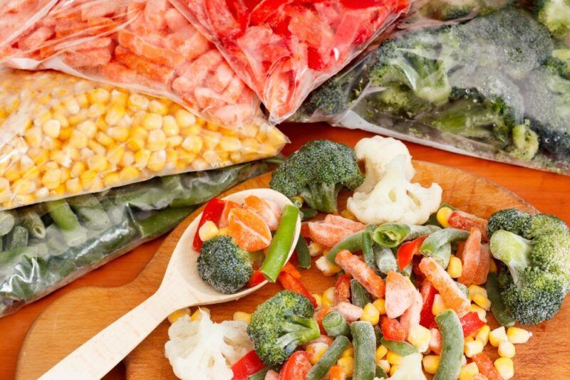 Bags of frozen vegetables on a table, half surrounding a board with loose frozen vegetables