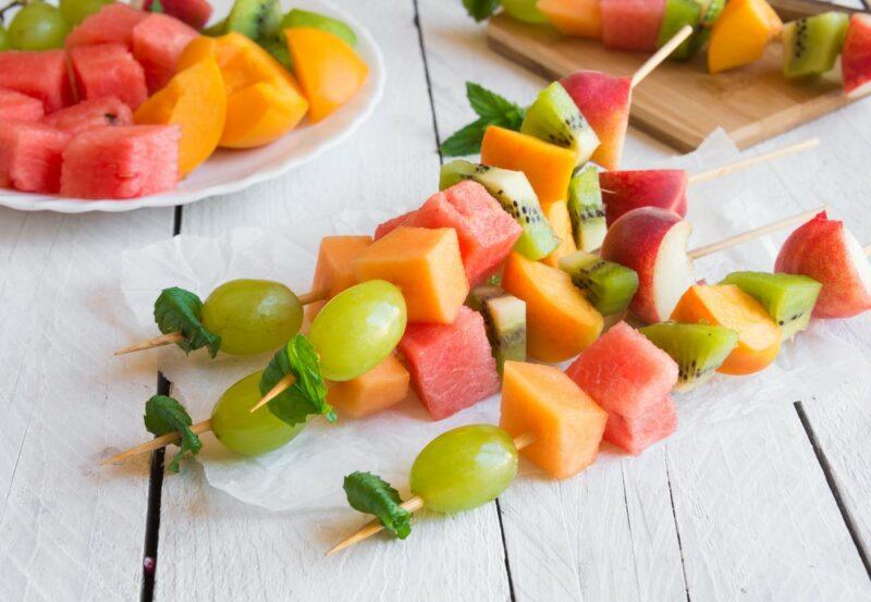 Four fruit kebabs on a white wooden table, with a plate of fruit in the background