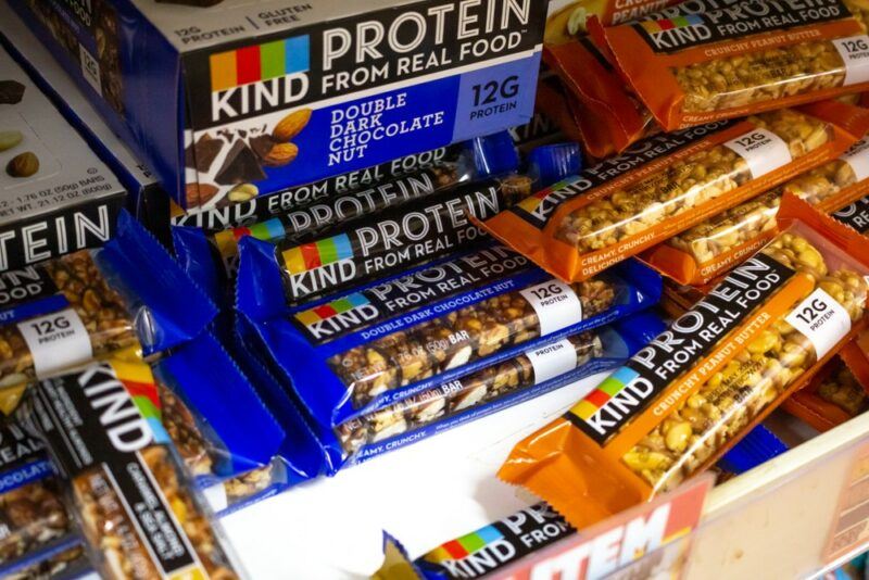 A box of Kind protein bars and many individual bars on the table and in piles