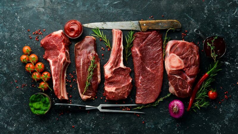 Various raw red meats on a black slate table, next to a knife, a carving form, and some tomatoes