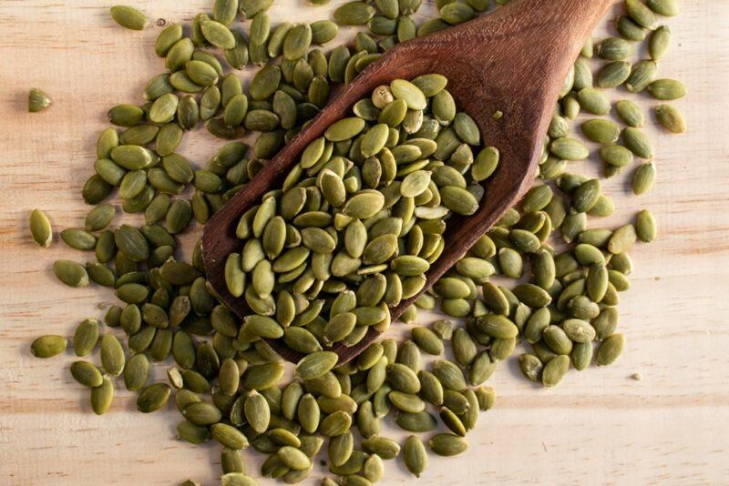 A light wooden table containing many pumpkin seeds and a dark wooden spoon