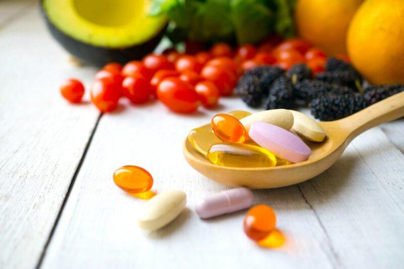 A white wooden table with a selection of fresh fruit, in front of which is a spoon of supplements and a few supplements on the table