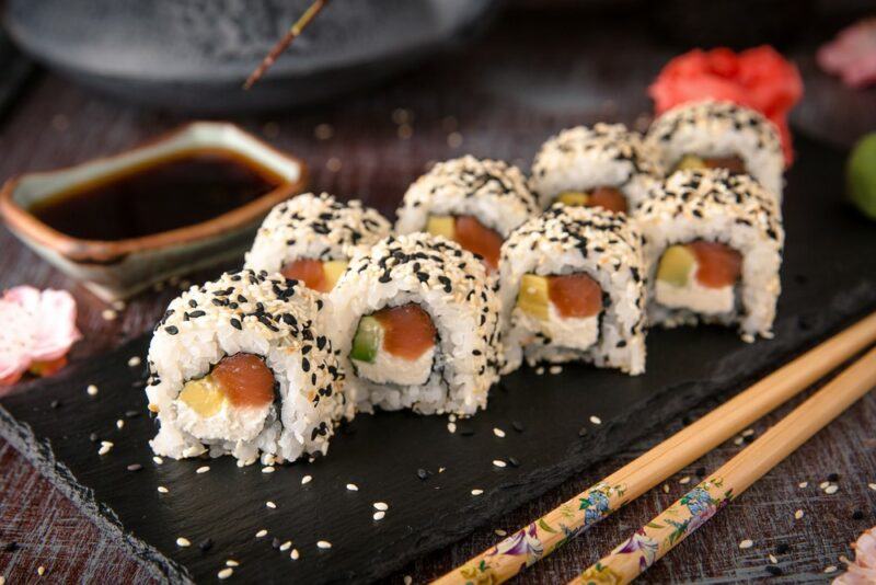 A black slate with sushi that contains tuna, cream cheese, and avocado, with black sesame seeds on top