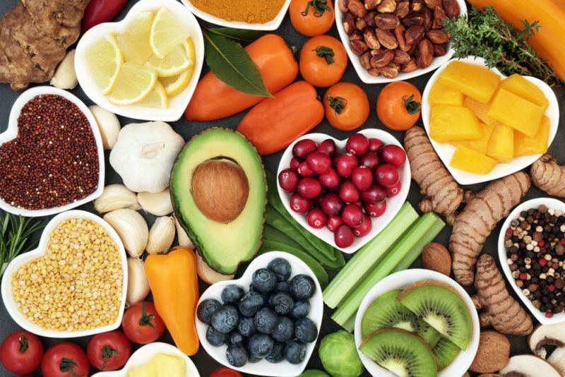 A large selection of healthy whole foods on a table, many of which are in white heart shaped dishes. The foods include kiwifruit, avocado, carrots, legumes, and fruit