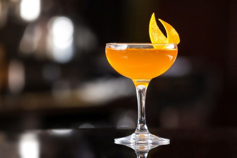 A coupe glass containing a sidecar cocktail with an orange twist, in front of a darkened bar