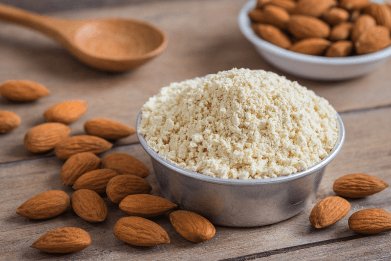 A wooden table with a metal bowl of almond flour, a small bowl of almonds, and almonds scattered on the table