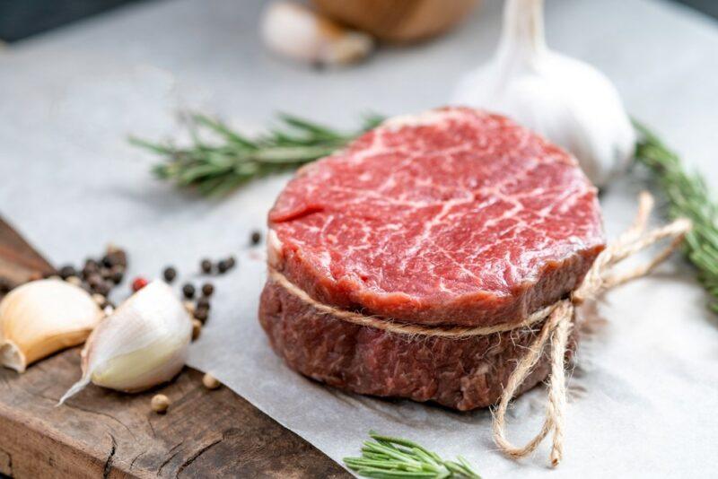 A piece of filet mignon that is wrapped with string, with garlic, rosemary, and black pepper surrounding it