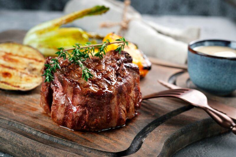 A wooden board with a single cooked filet mignon steak next to some seasoning ingredients, a knife and a fork