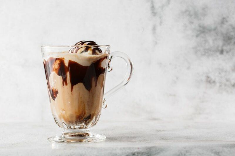 A large glass containing a chocolate peanut butter cup milkshake or cocktail with ice cream, on a gray table in front of a gray wall