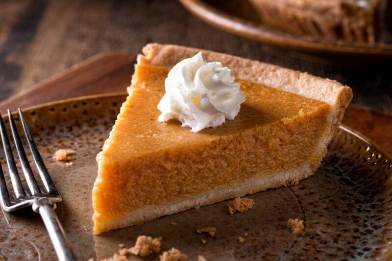 A brown plate containing a single piece of pumpkin pie with cream, next to a fork